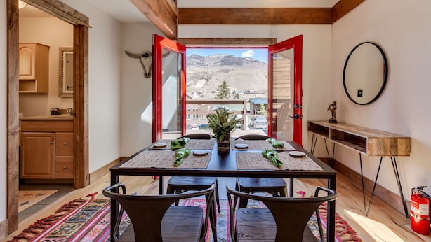 Un coin repas avec une table dressée pour quatre personnes, un plafond en bois et une vue sur les montagnes par des portes ouvertes.