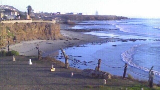 Eine Küstenlandschaft mit Kiesstrand, einer sanften Grasflanke und einem Gebäude in der Ferne.