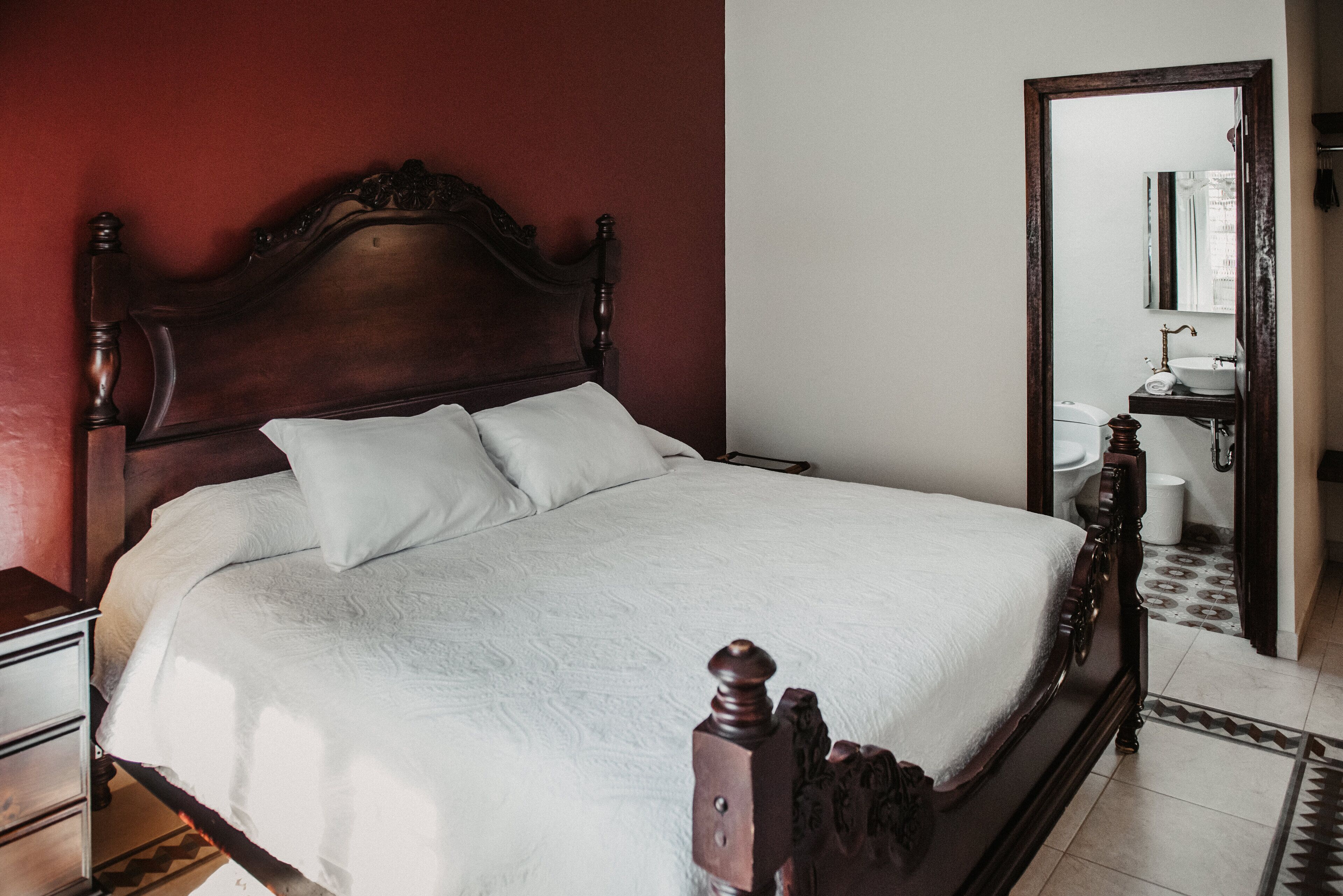 A neatly made bed with a wooden headboard, a bedside table, and a mirror above the sink in a bathroom.