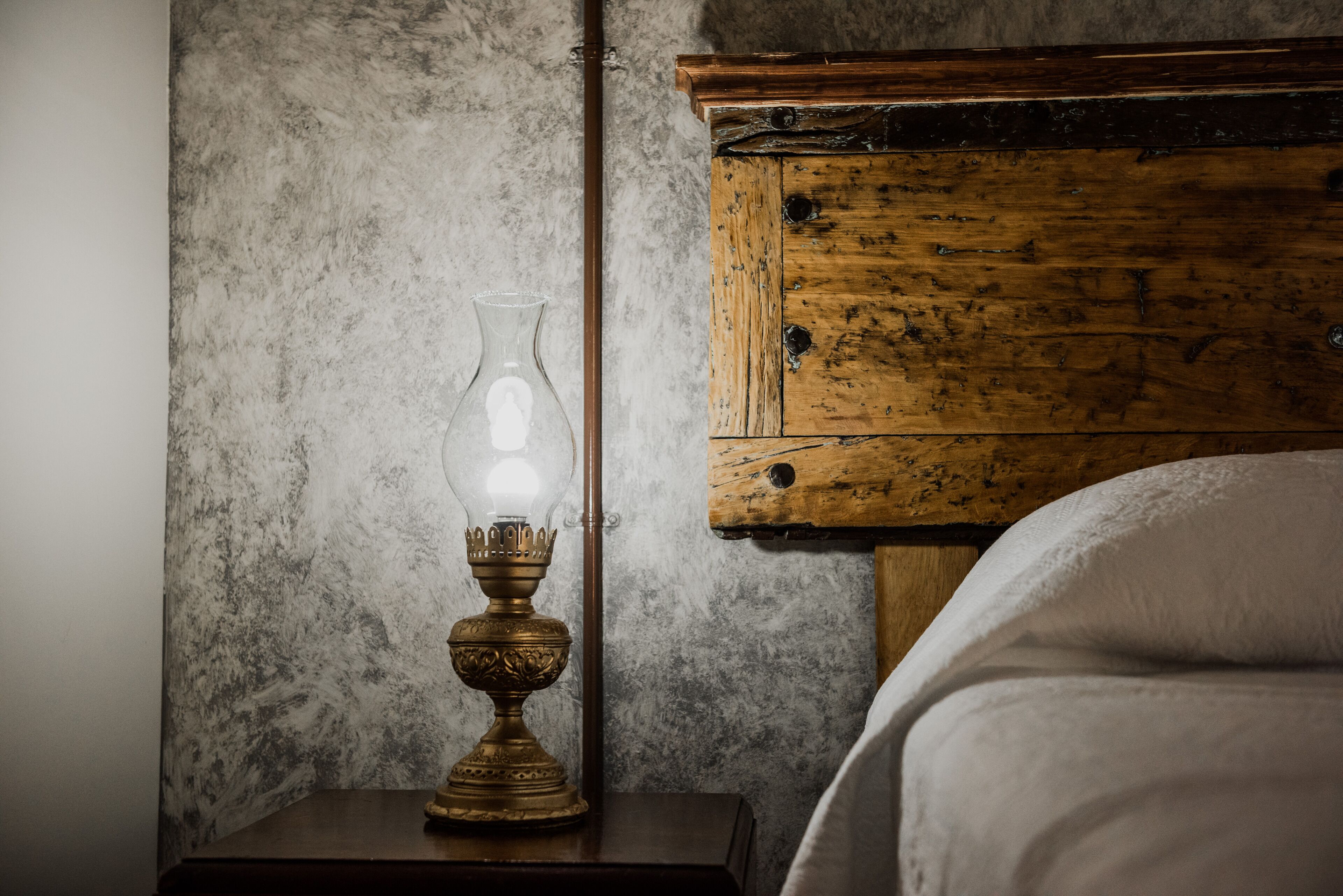A wooden nightstand with a clear glass lamp and a white bed in the background.