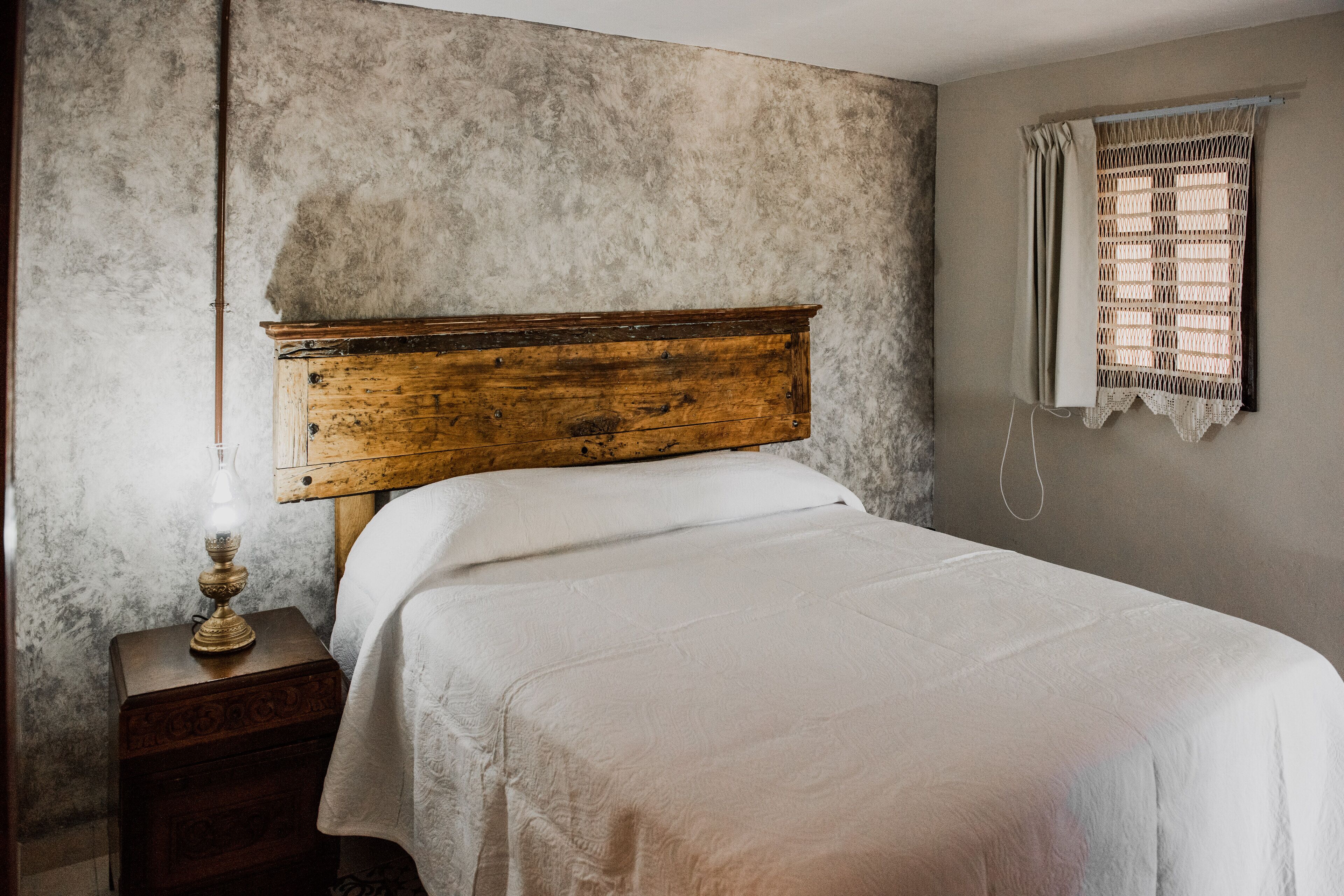 A bedroom with a wooden headboard, a bedside table with a lamp, and a window with a woven blind.