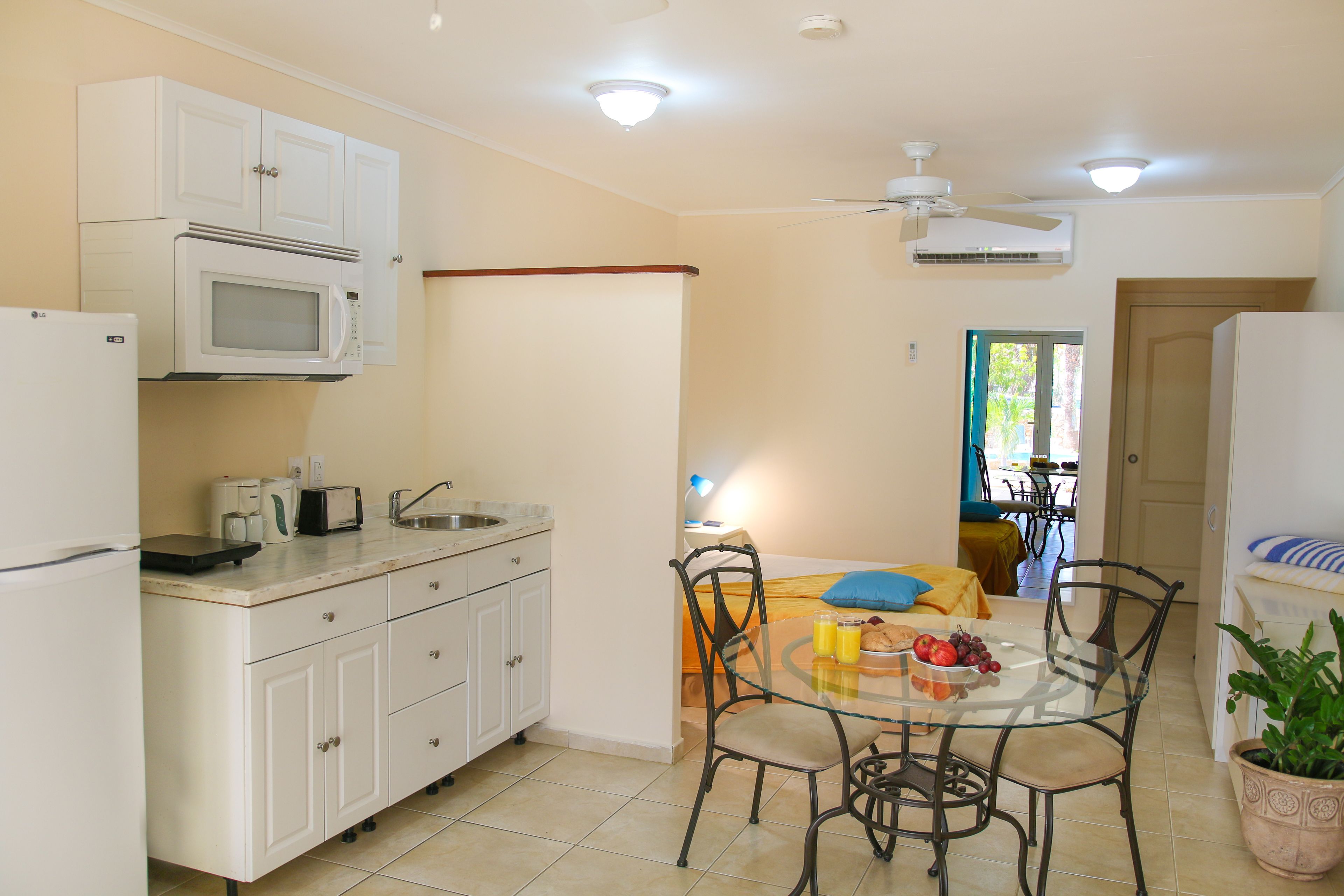 A compact kitchen with white cabinets, a microwave, and a refrigerator. There is a dining area with a glass table and chairs, and a potted plant.