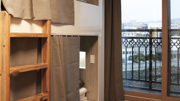 A room with a bunk bed, a wooden ladder, a window with a view of buildings, and a balcony with a railing. - Bed Radio Dongmoon - Hostel (Jeju City)
