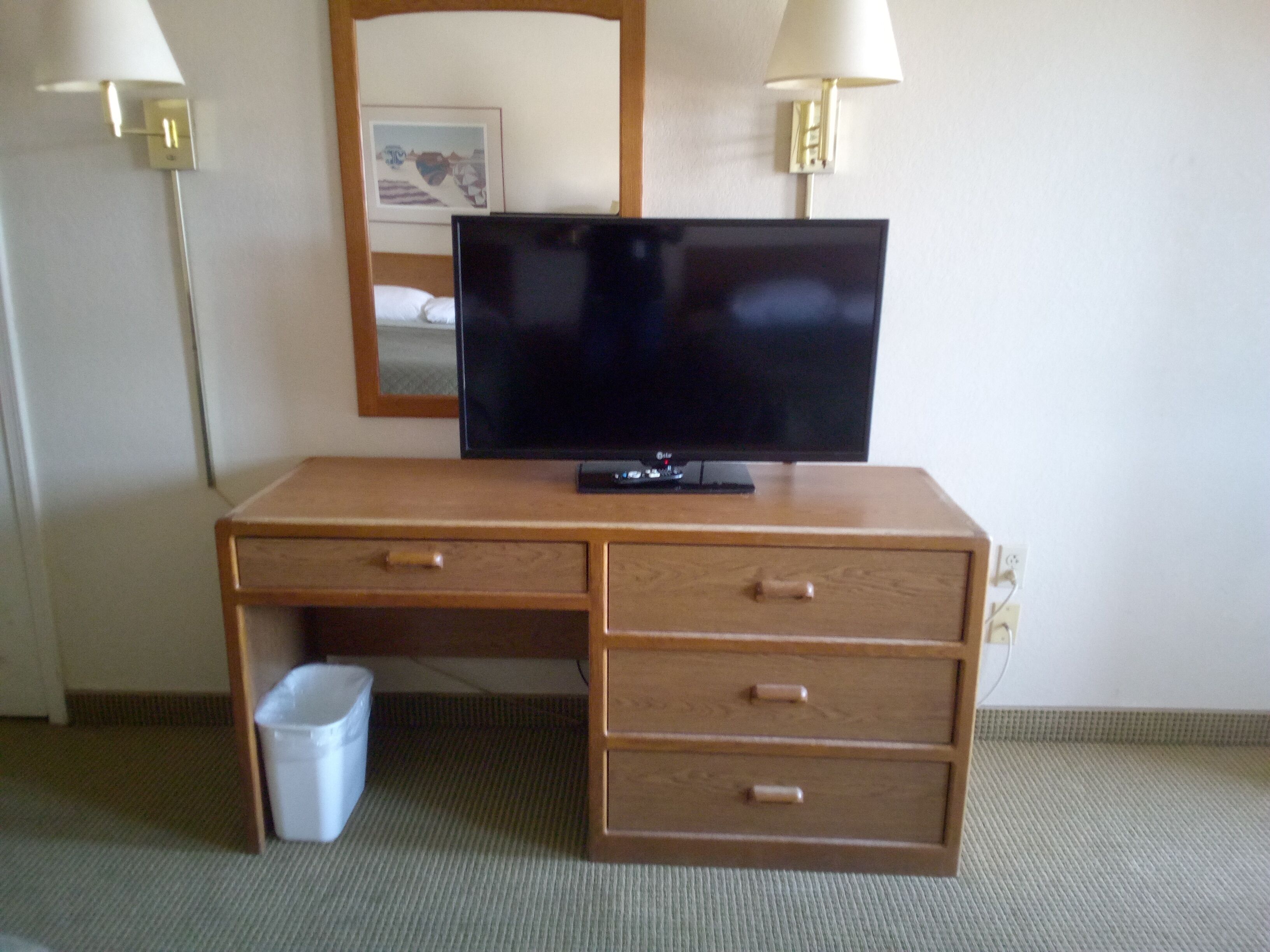 A wooden desk with a flat-screen TV, two lamps, a trash bin, and a framed picture on the wall.