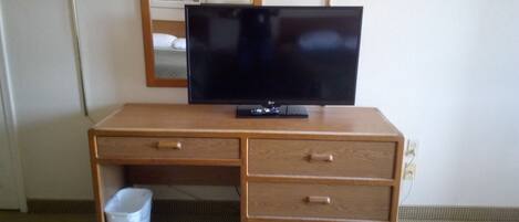 A wooden desk with a flat-screen TV, two lamps, a trash bin, and a framed picture on the wall.