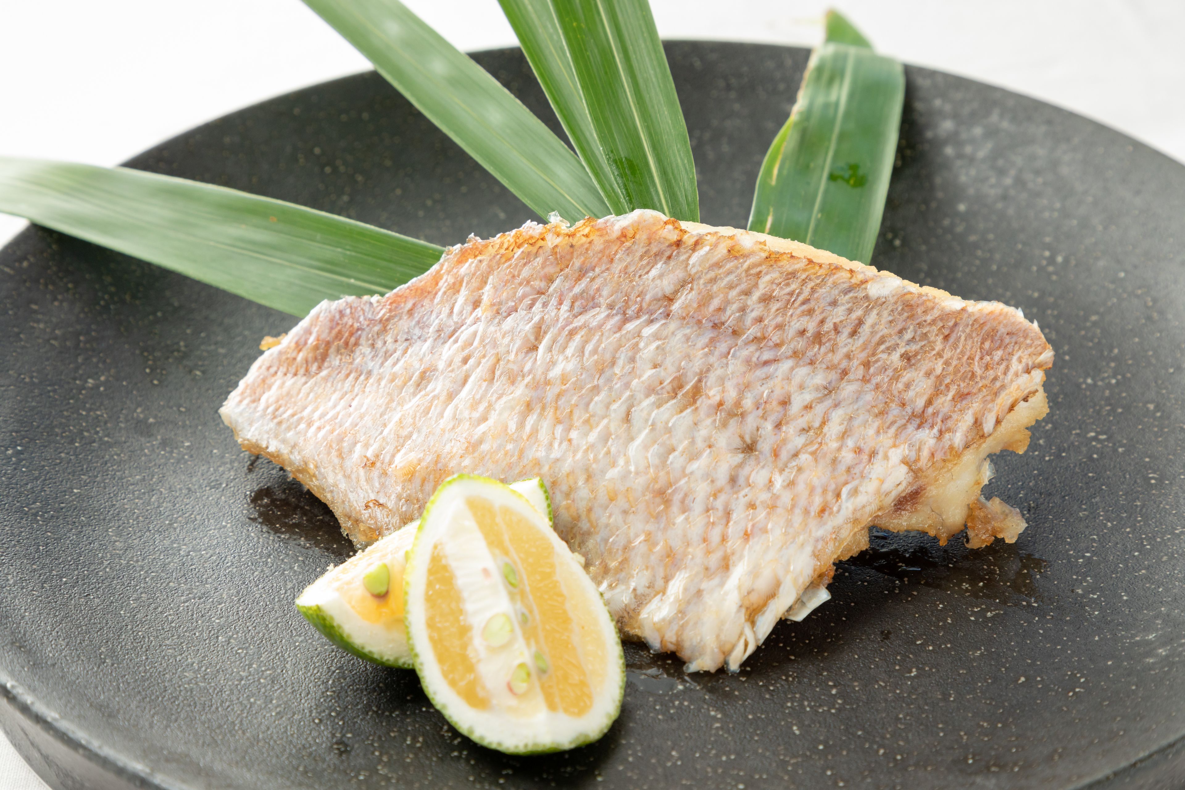 A piece of grilled fish on a black plate with a slice of lime and green leaves.