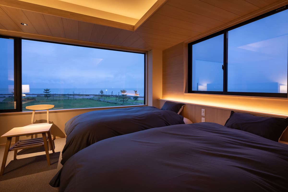 A bedroom with two beds, a small table, and large windows with a view of the ocean and trees.