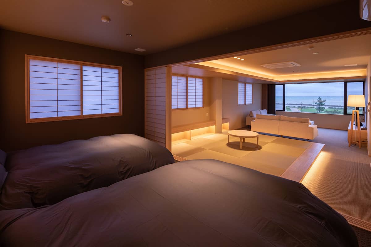 A modern hotel room with two beds, a small sitting area, and a view of the sea.