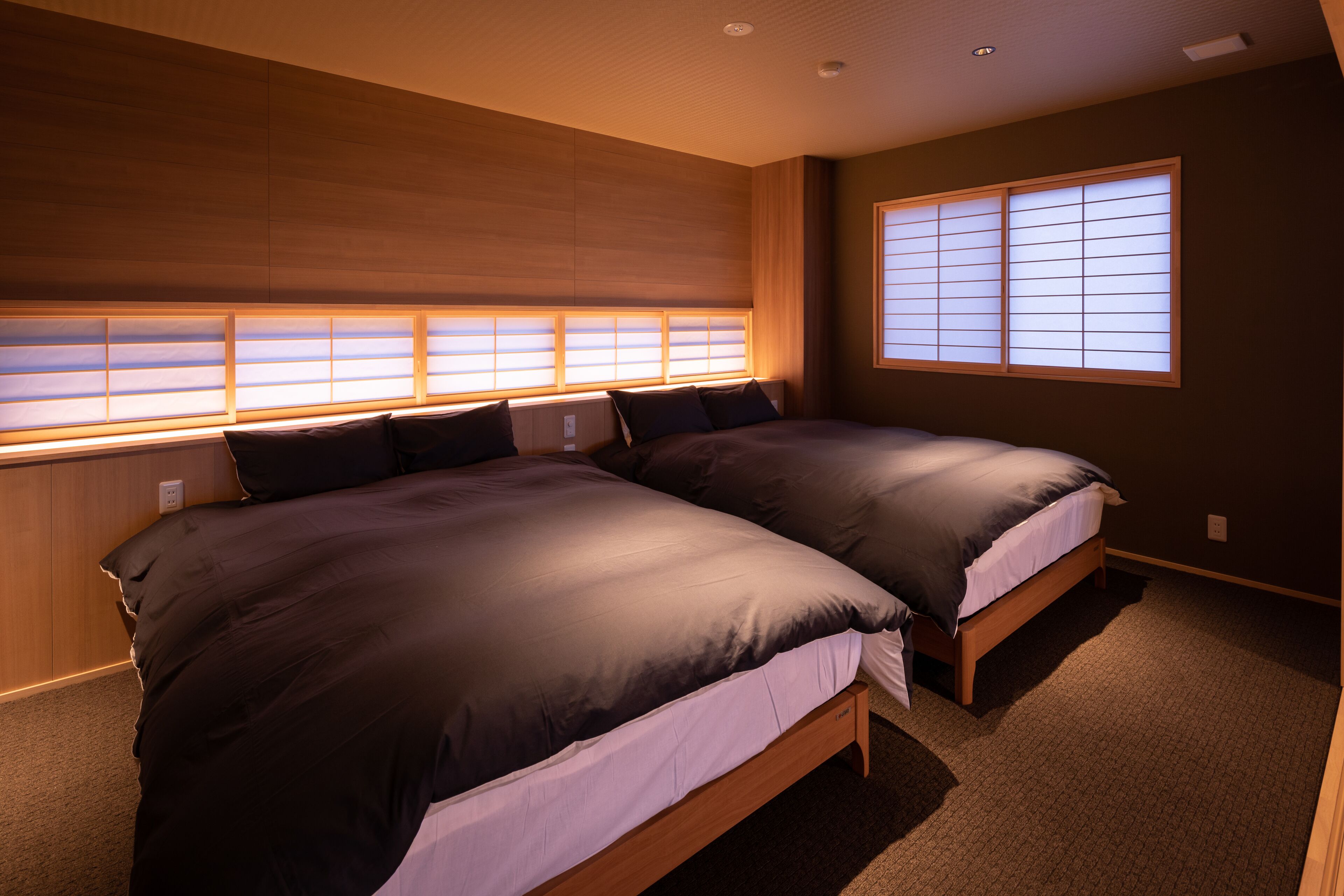Ein Schlafzimmer mit zwei Betten, einem Holzkopfstück, großen Fenstern mit Jalousien und Teppichboden.