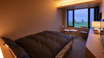 Quarto de hotel com uma cama grande, uma escrivaninha com cadeira, uma mesinha e vista para o exterior através de amplas janelas.