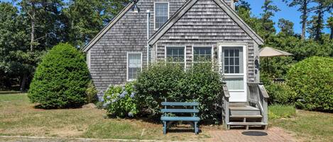 A two-story wooden house with a porch, surrounded by trees and shrubs.