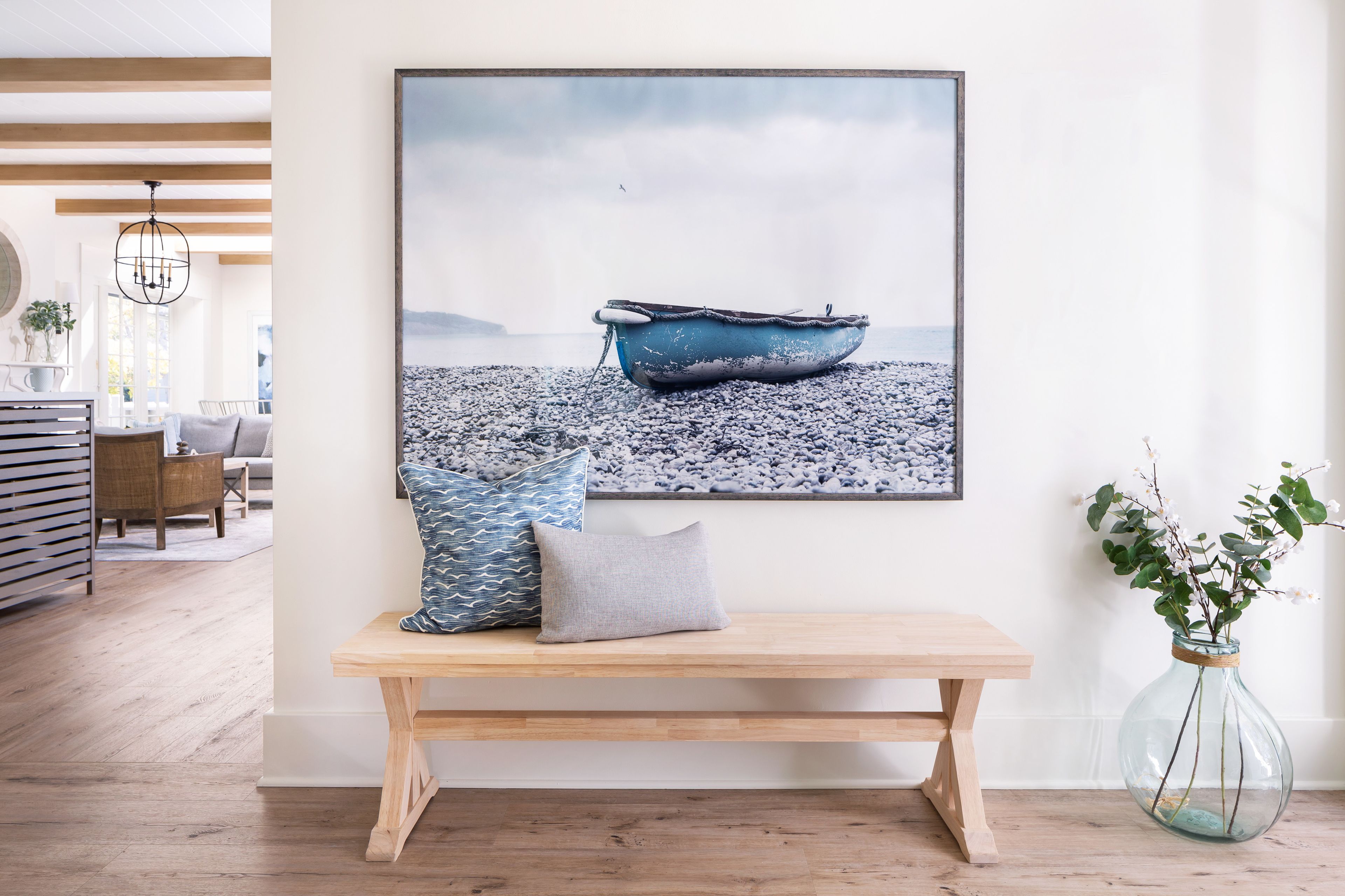 A room with a wooden bench, a framed picture of a boat on water, and a vase with plants.