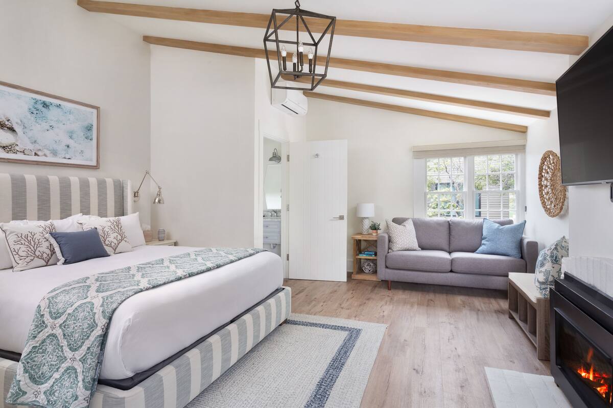 A bedroom with a bed, bedside table, and a nightstand. There is a sofa and a fireplace in the living area.