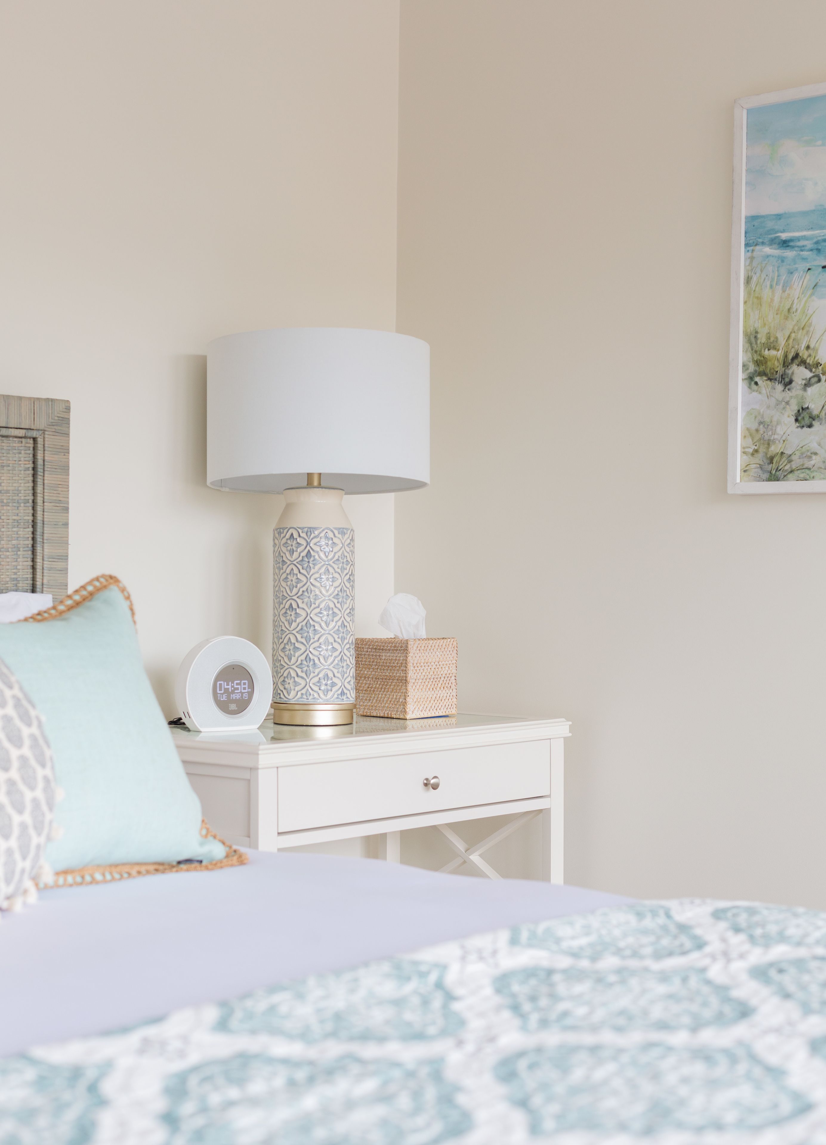 A neatly arranged bedroom with a bed, a bedside table with a lamp, a clock, and a framed picture on the wall.