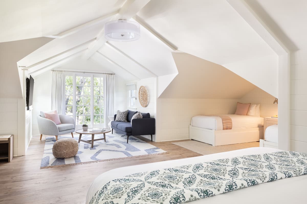 A spacious loft bedroom with a large bed, a sitting area with a sofa and armchair, and a view of the outdoors through a large window.