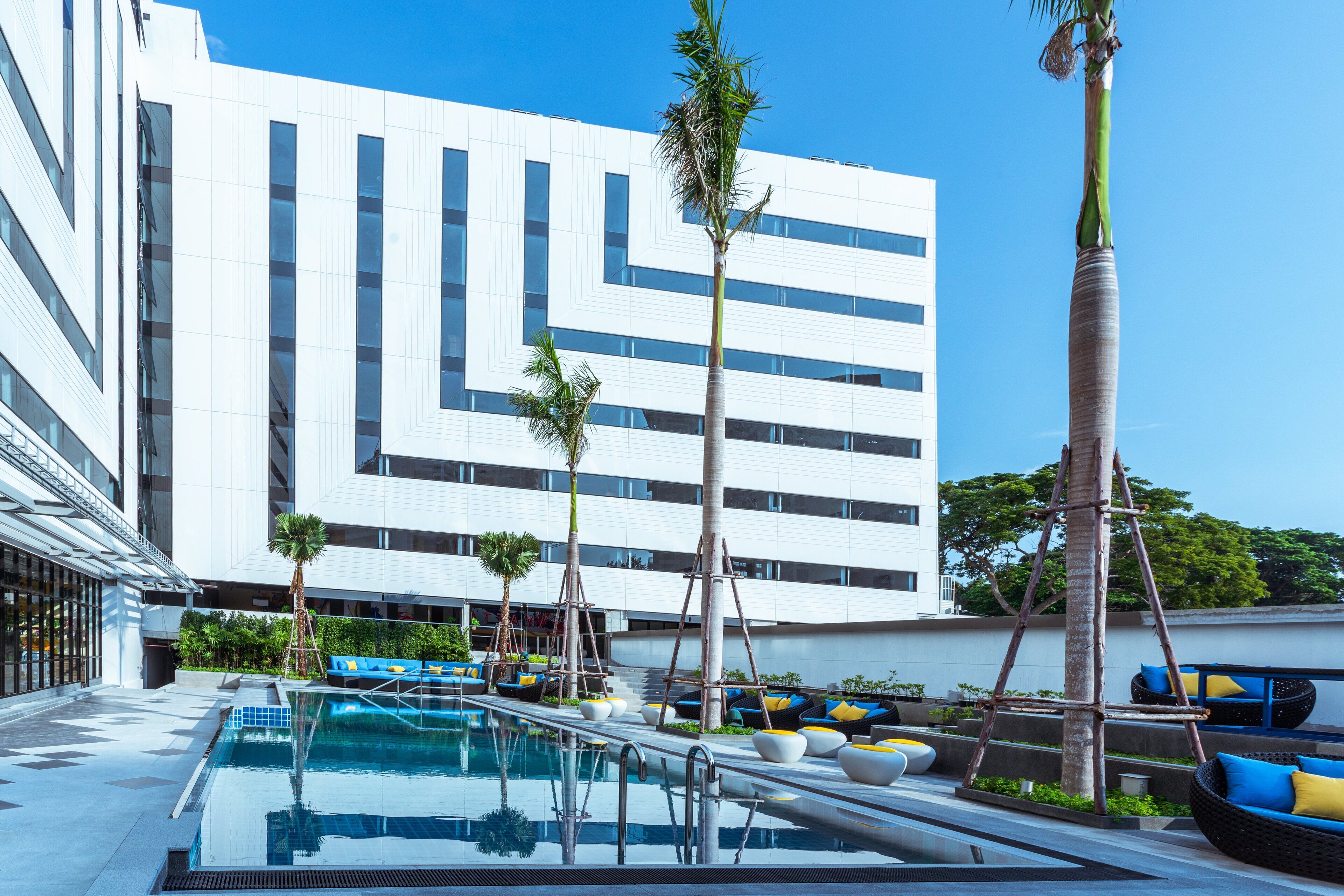 Un hotel moderno con zona de piscina, mobiliario exterior y un edificio grande de fachada geométrica llamativa.