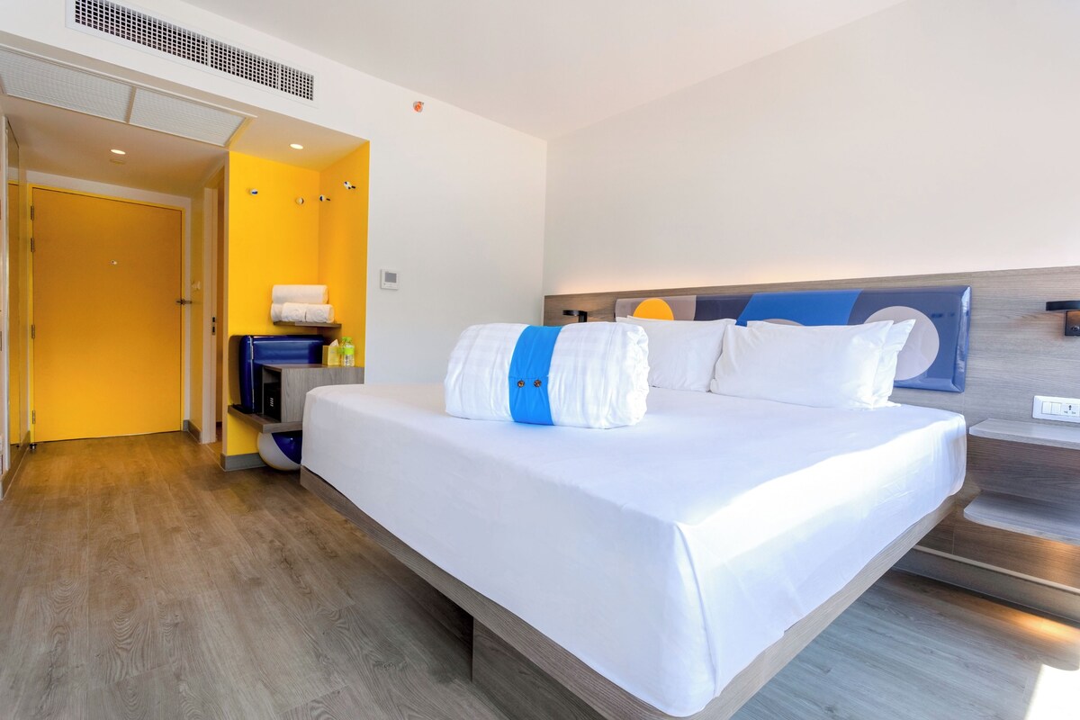 A hotel room with a large bed, a wooden floor, a yellow wall, and a door leading to a bathroom.