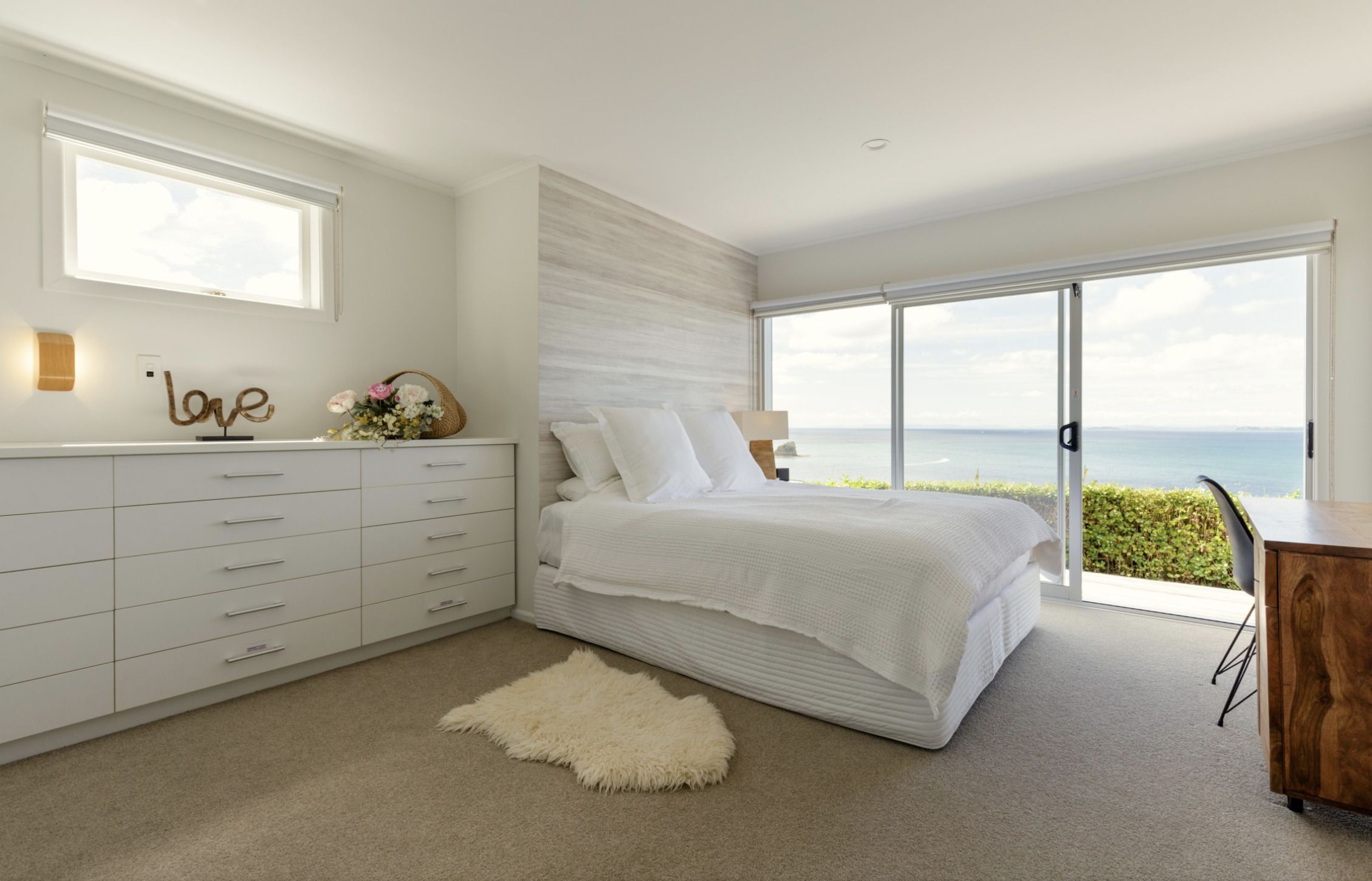A modern bedroom with a large bed, a white dresser, a fluffy rug, and a view of the sea through a sliding glass door.