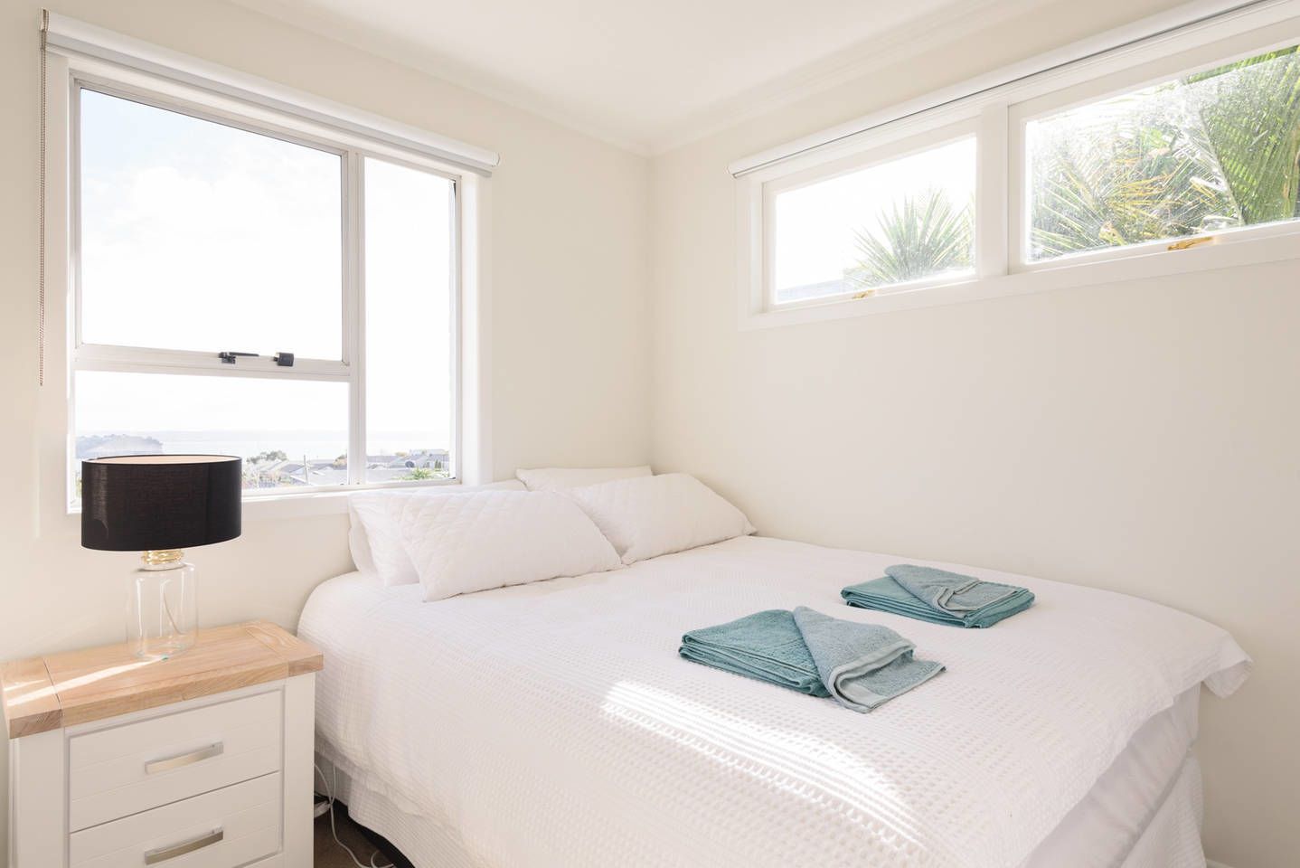 A bedroom with a bed, two windows, a bedside table with a lamp, and a view of the outdoors.