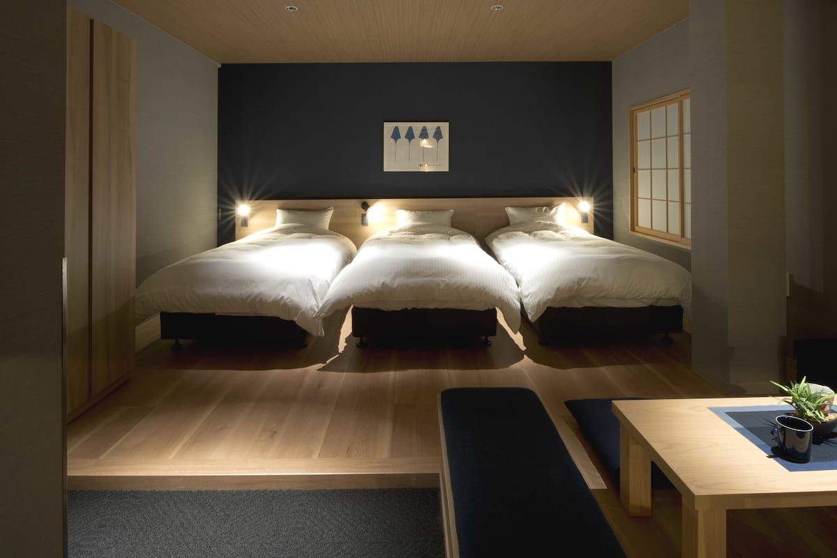 Een moderne slaapkamer met drie bedden, een houten vloer, een klein tafeltje met een plant en een aan de muur hangend schilderij.
