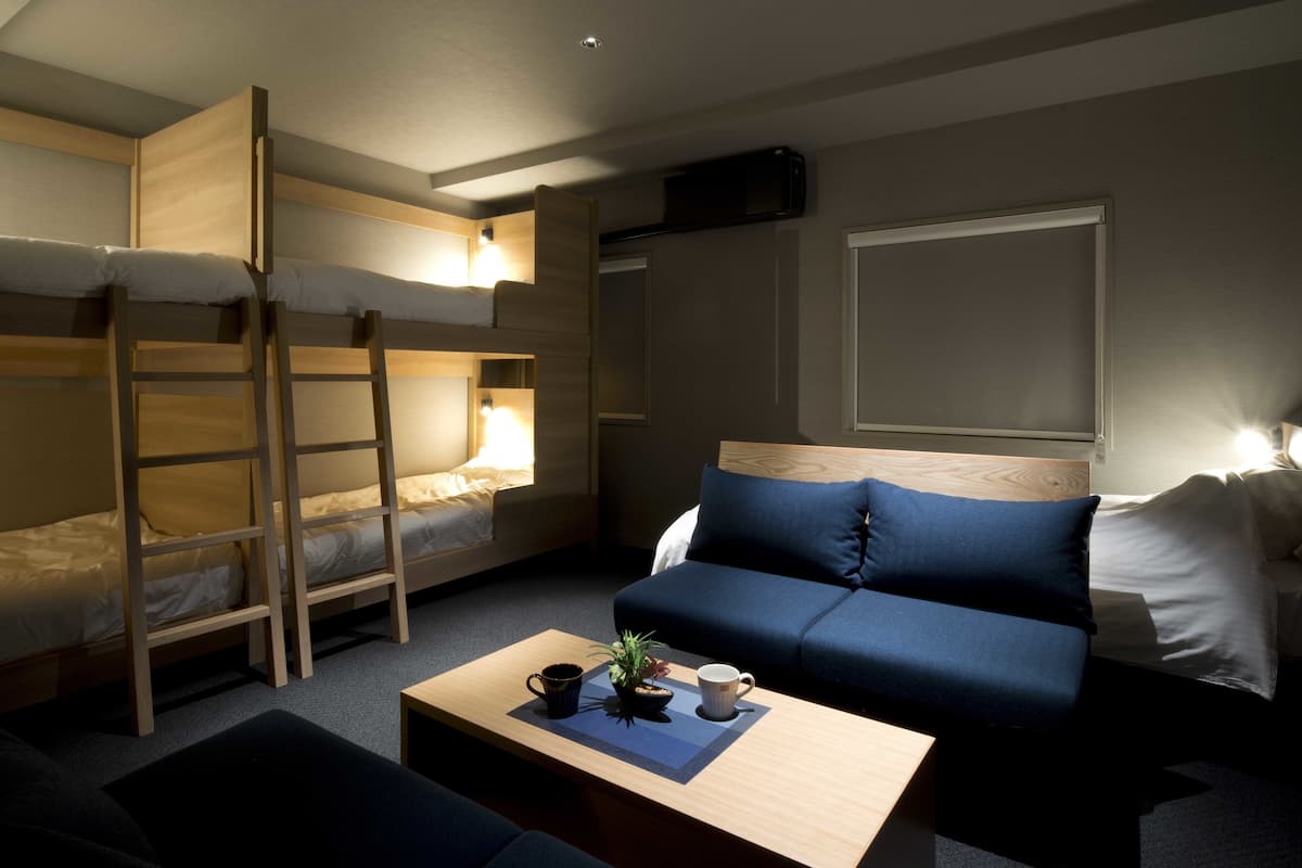 Een kleine hotelkamer met stapelbedden, een slaapbank, een bijzettafeltje met een plant en beker, en een televisie aan de muur.