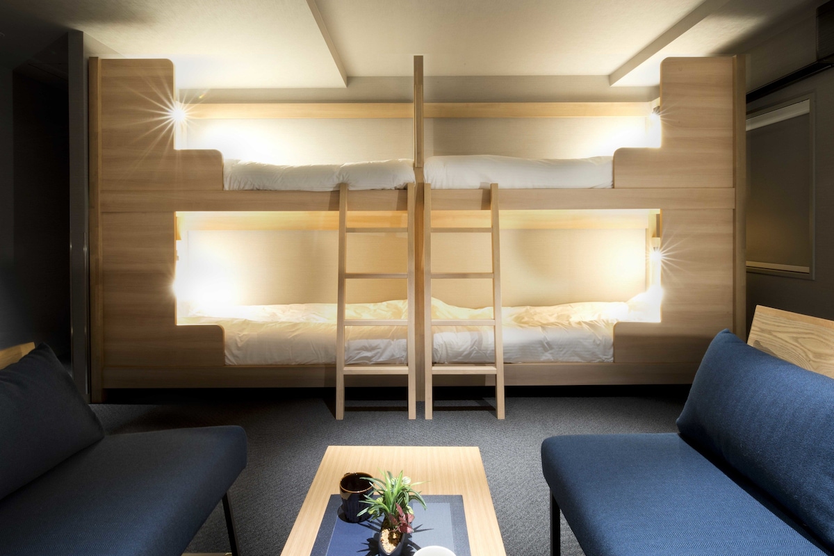 Een slaapkamer met een stapelbed, twee blauwe slaapbanken, een kleine tafel met een plant en een lichtbron boven de bedden.