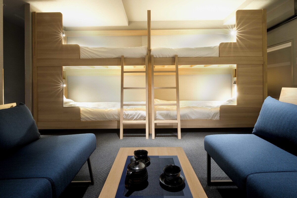 Een kamer met stapelbedden, een ladder, twee blauwe banksofa's en een tafel met theeserviesjes.