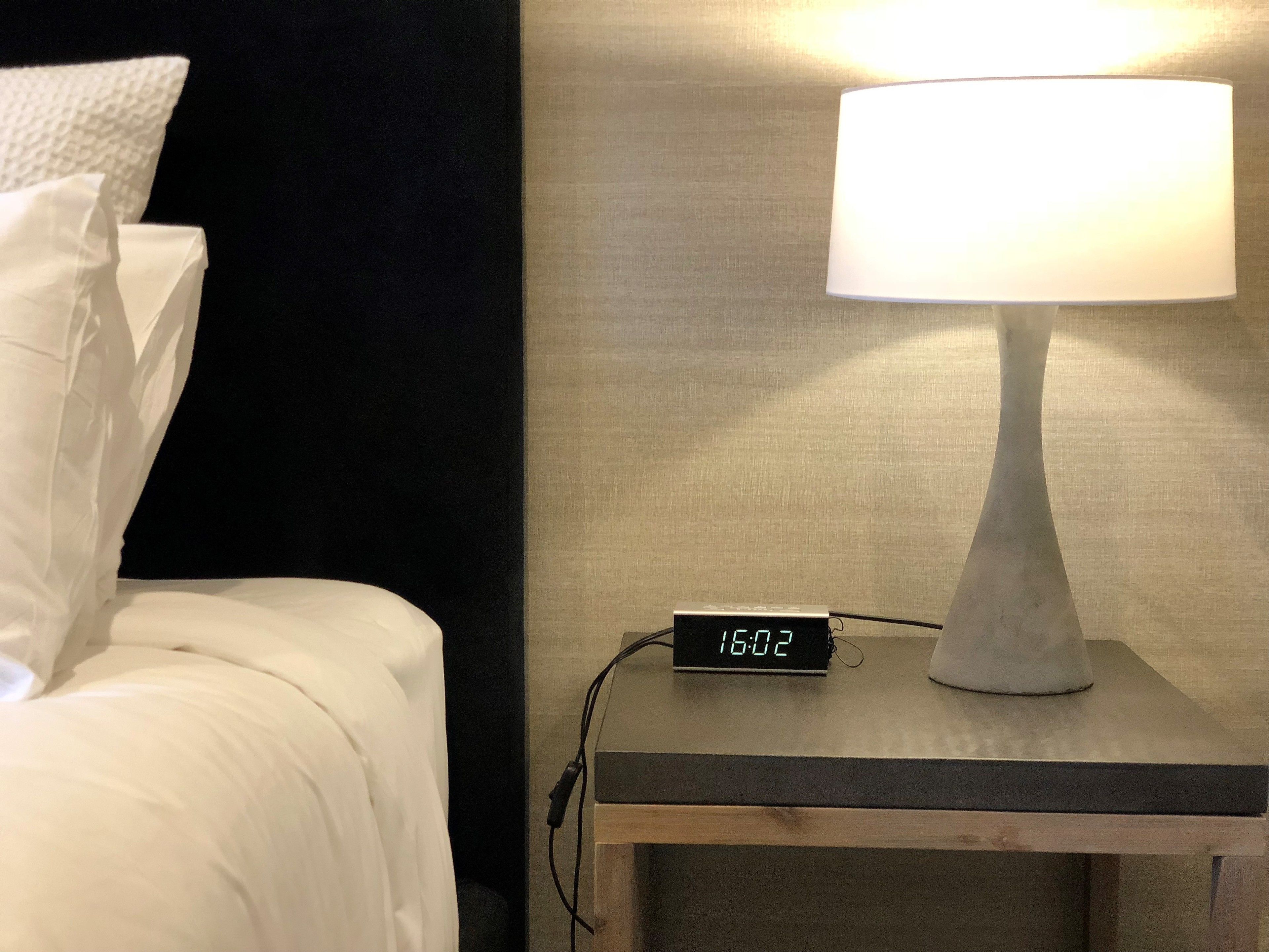 Ein Bett mit weißen Leinen und Kissen, ein Nachttisch mit Lampe und einer digitalen Uhr, die 16:02 anzeigt, sowie eine strukturierte Wand im Hintergrund.
