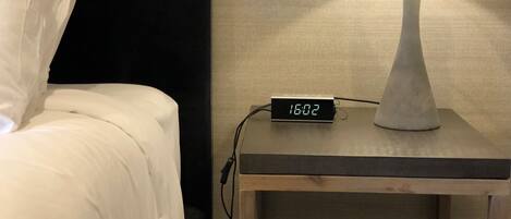 Ein Bett mit weißen Leinen und Kissen, ein Nachttisch mit Lampe und einer digitalen Uhr, die 16:02 anzeigt, sowie eine strukturierte Wand im Hintergrund.