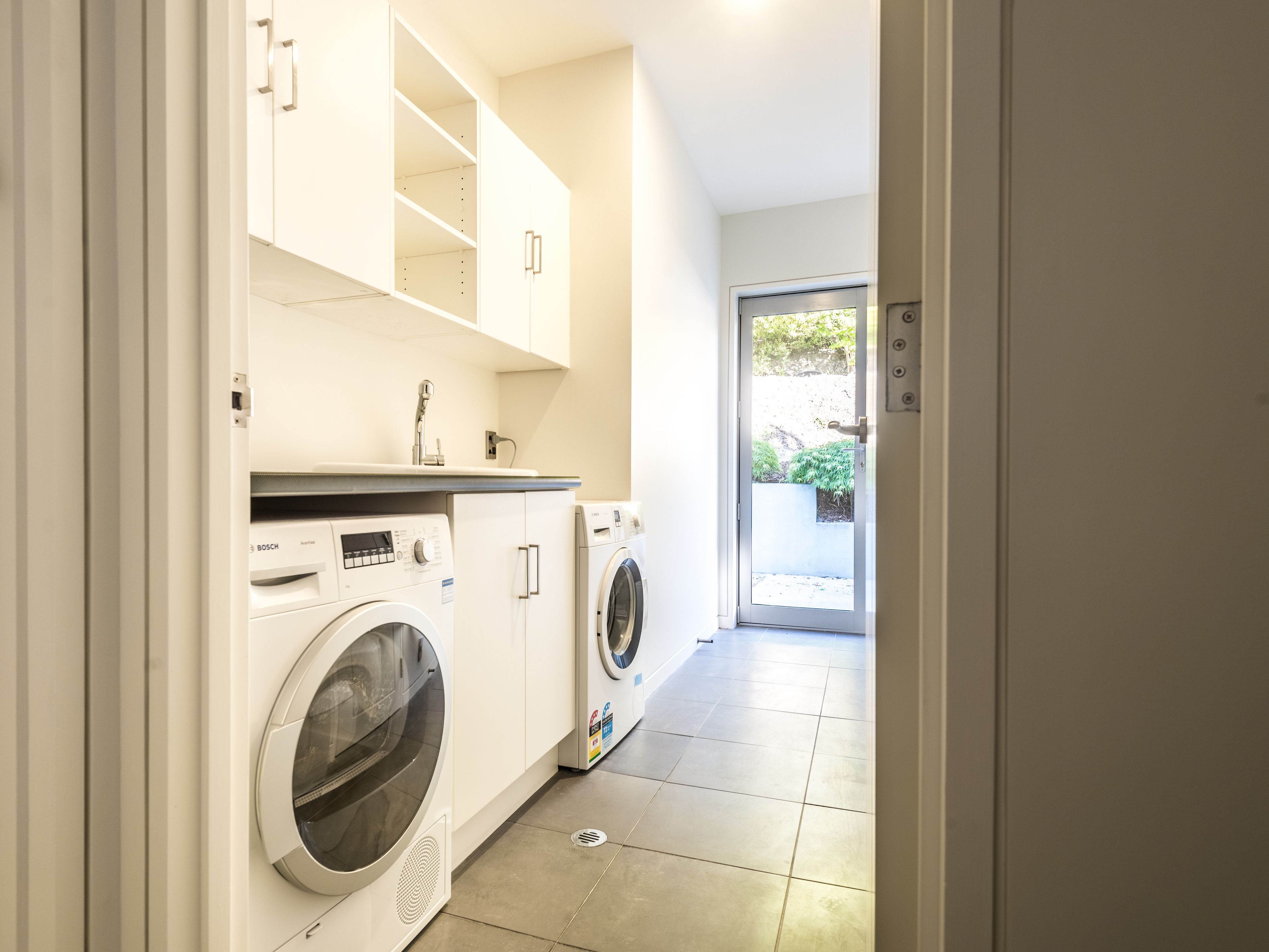 Una sala de lavandería con lavadora y secadora Bosch, armarios blancos y piso de baldosas.