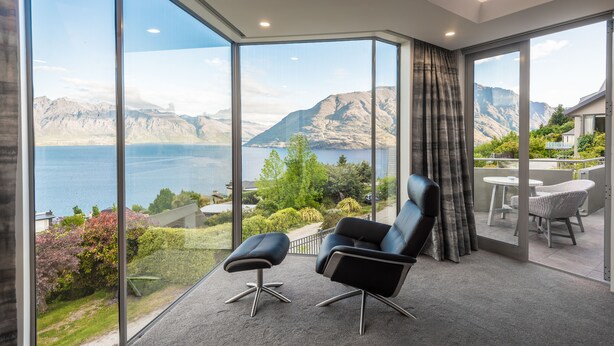Una habitación moderna con amplios ventanales de vidrio, un sillón de cuero negro, un pequeño reposapiés y vistas a las montañas y al agua.