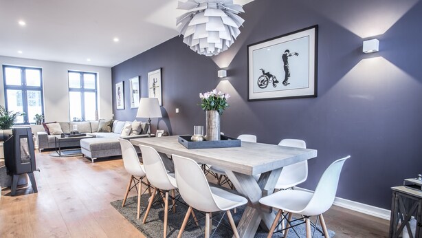 Un salon moderne avec un canapé gris, une table à manger entourée de chaises blanches et une grande fenêtre.