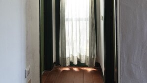 Un corridoio stretto con pavimento piastrellato, una finestra con tende leggere e uno specchio fissato alla parete.