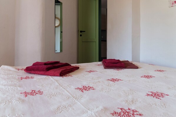 Un lit recouvert d’une couverture blanche ornée de motifs floraux rouges, deux serviettes rouges pliées, une porte verte et un miroir accroché au mur.