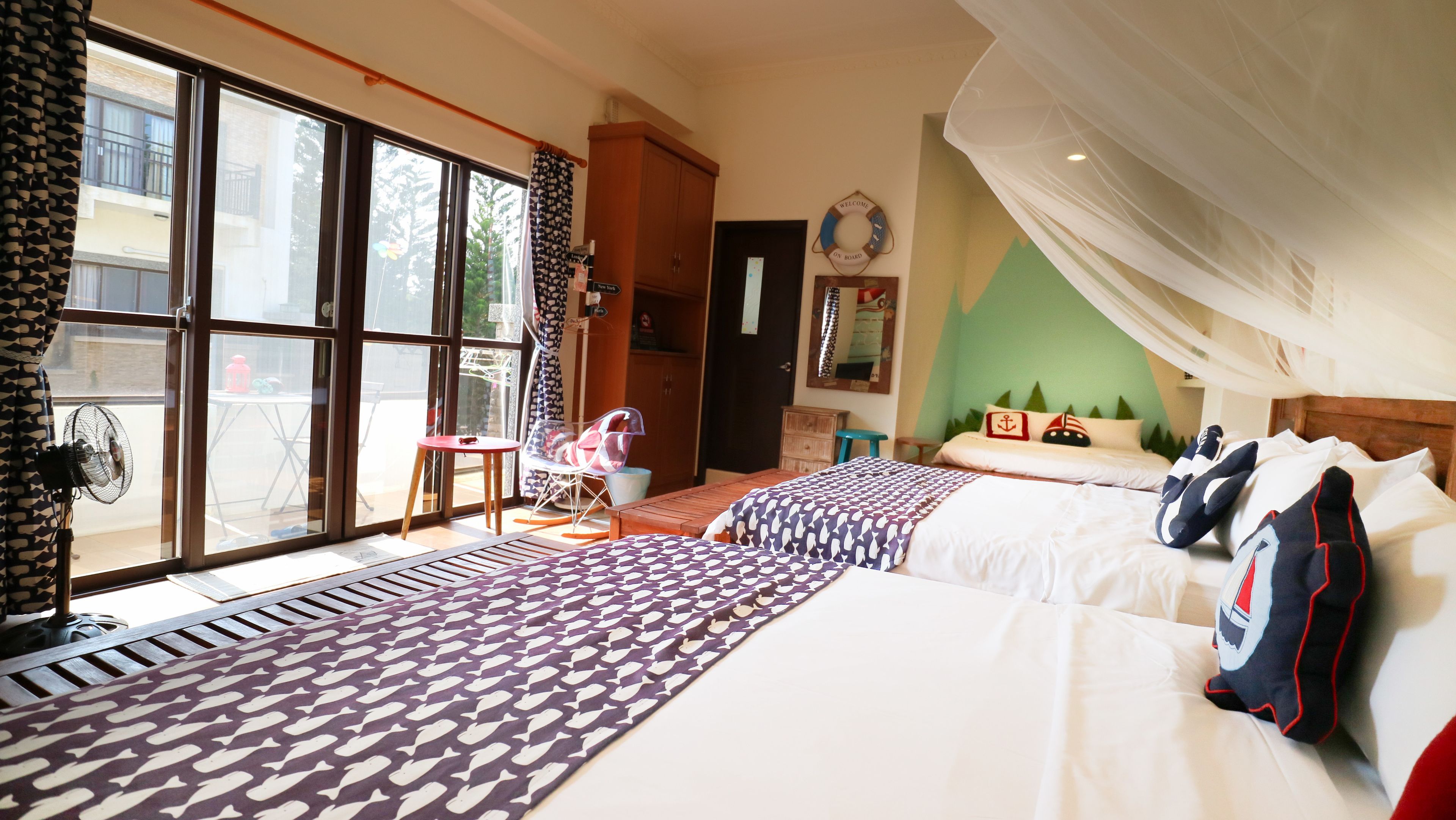 A hotel room with two beds, a large window, a fan, and a view of the outside balcony.