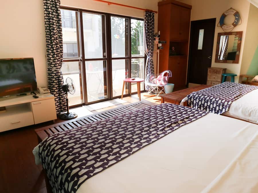Hotellihuone, jossa on kaksi sänkyä, televisio, tuuletin, ikkuna verhoilla ja pieni pöytä.