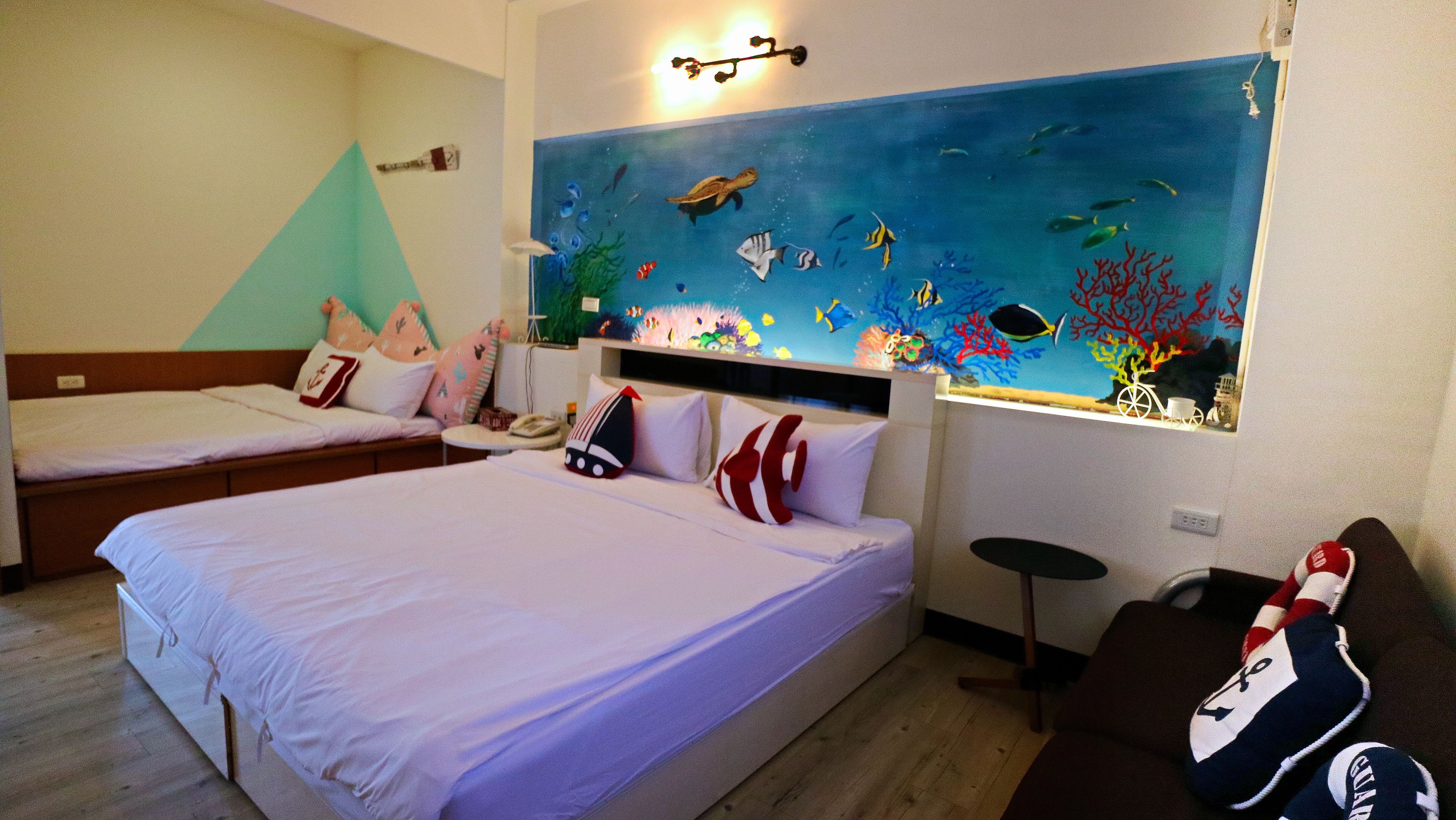 A hotel room with two beds, a large wall mural of an underwater scene, and a small table with a chair.