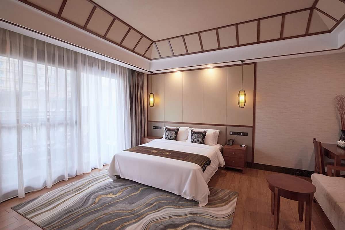 Ein Hotelzimmer mit einem großen Bett, einem Schreibtisch und einem Stuhl. Es gibt ein Fenster mit Vorhängen, einen Teppich und eine Wandleuchte.