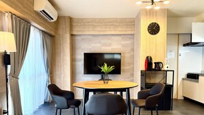 A modern room with a round wooden table, four black chairs, a flat-screen TV, a clock, a kitchen area with a coffee maker, and a plant on the table. - Reve K 302 (Naha)