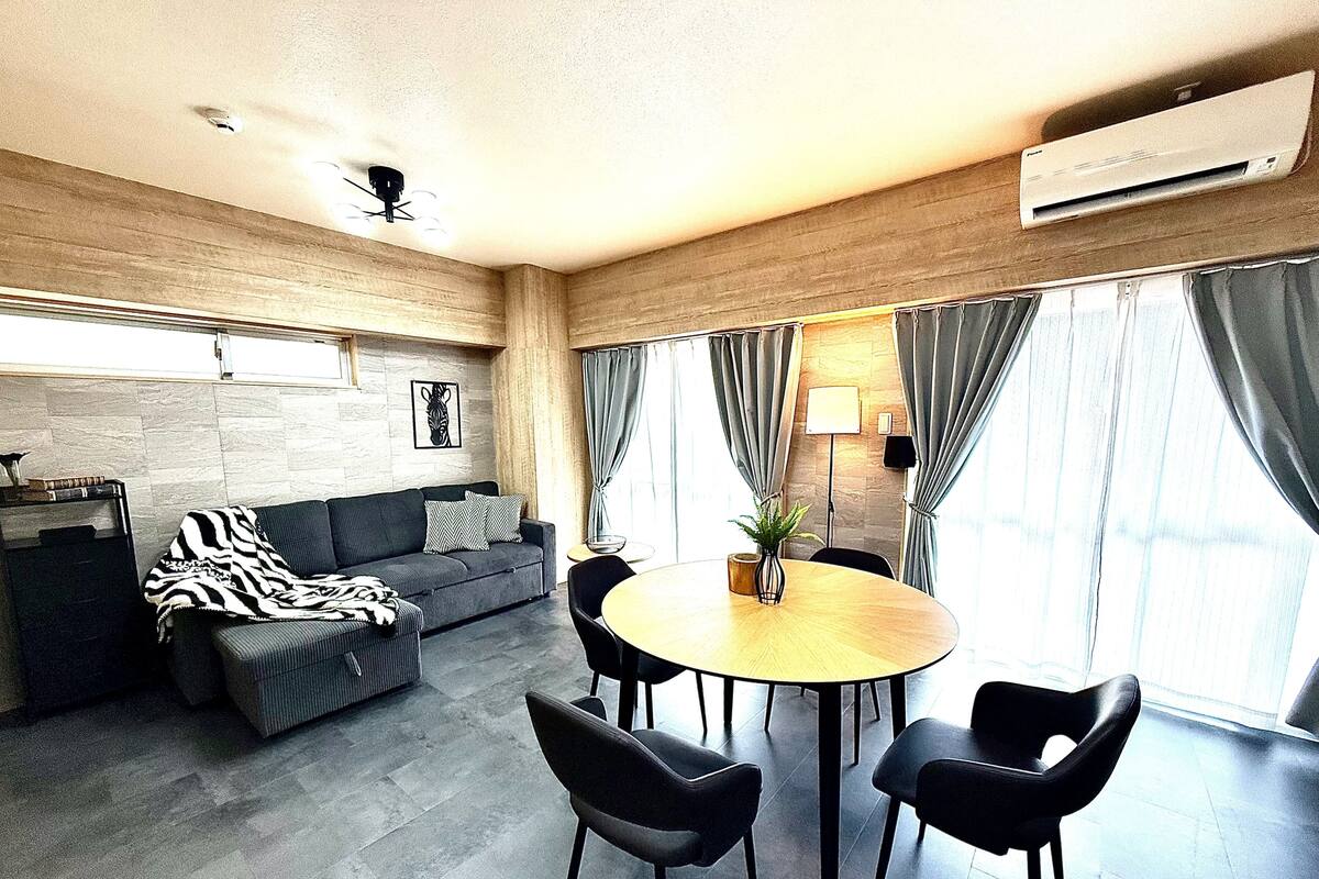 A modern living room with a sofa, a dining table with chairs, a ceiling fan, and a large window with curtains.