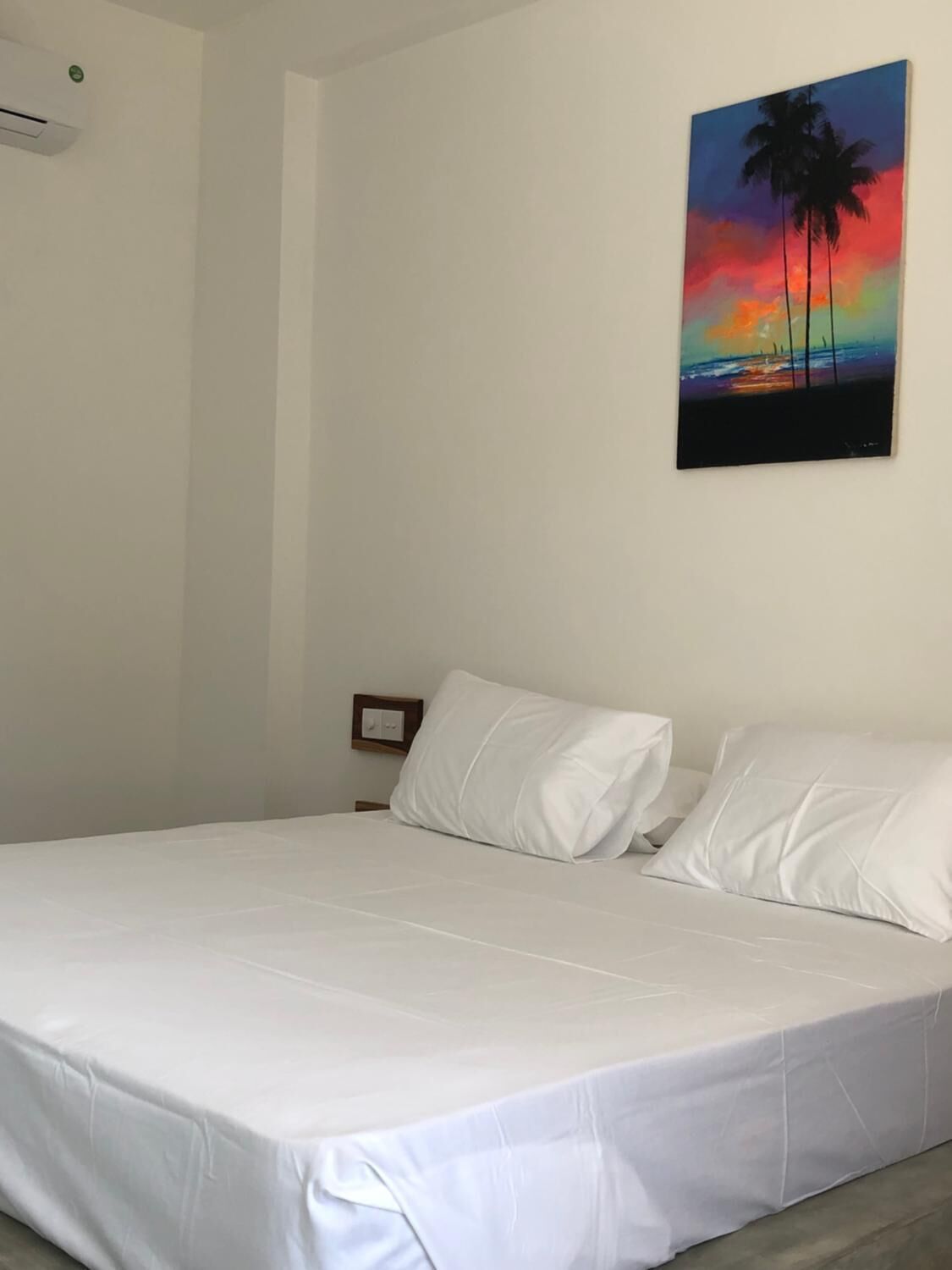 A bedroom with a bed, white bedding, a nightstand, a wall-mounted air conditioner, and a painting of a sunset with palm trees.