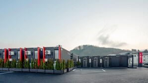A modern building with multiple air conditioning units, a parking area, and a gate leading to a building with numbered doors. - HOTEL R9 The Yard KANUMA NISHIMORO (Kanuma)