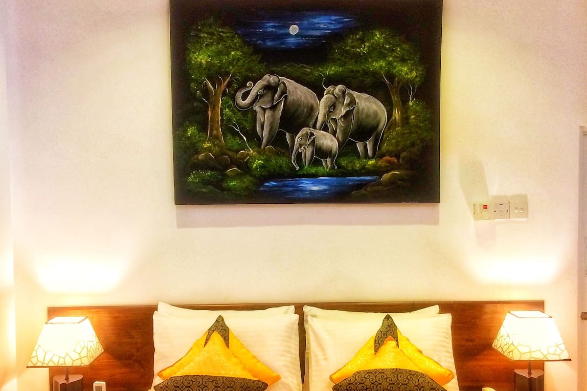 A hotel room with a bed, two bedside lamps, a nightstand, a painting of elephants, and a heart-shaped towel arrangement.