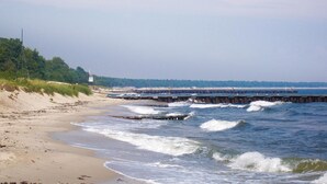 Una playa con un muelle de madera que se adentra en el agua, un faro al fondo y olas rompiendo sobre la orilla.