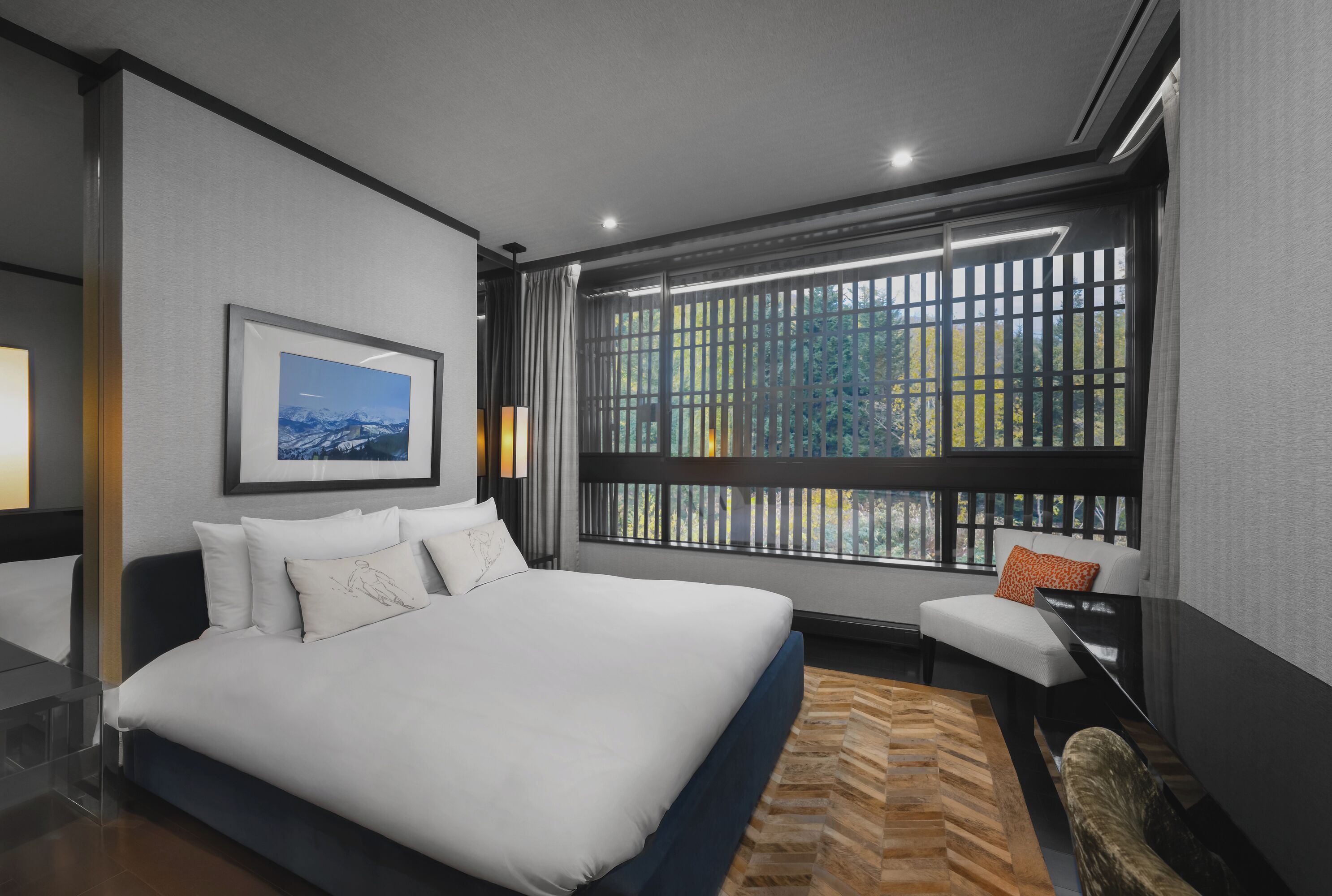 A modern hotel room with a large bed, a view of trees through a window, and a small seating area.