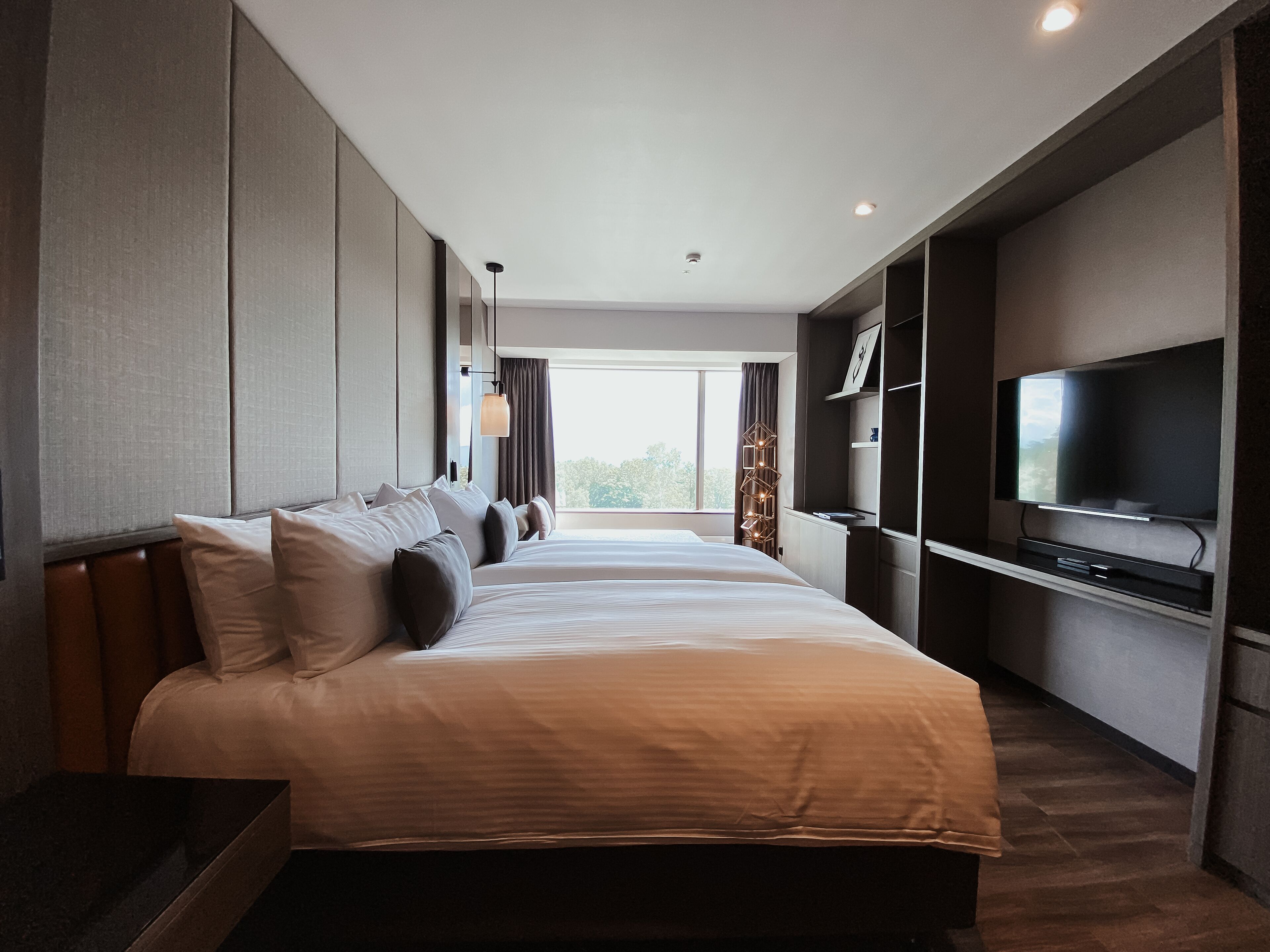 A modern hotel room with a large bed, a flat-screen TV mounted on the wall, and a view of the outdoors through the window.