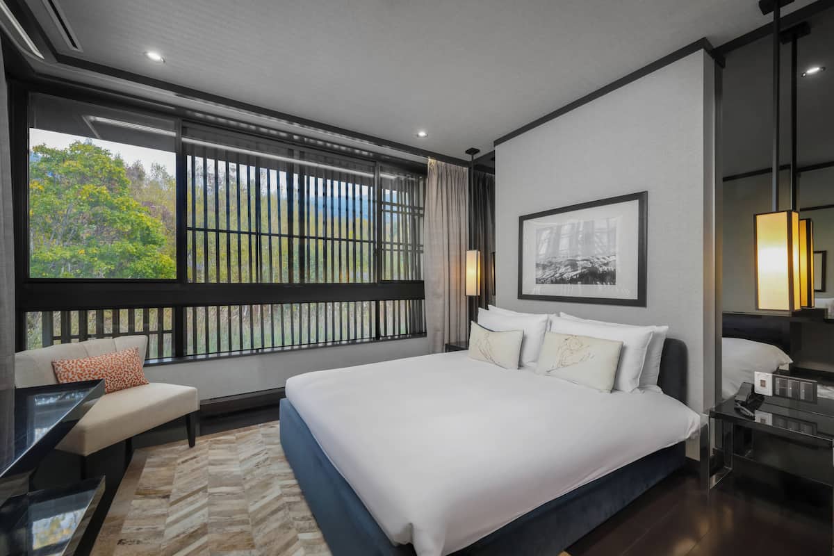 A modern hotel room with a large bed, a sofa, a desk, and a view of greenery through the window.