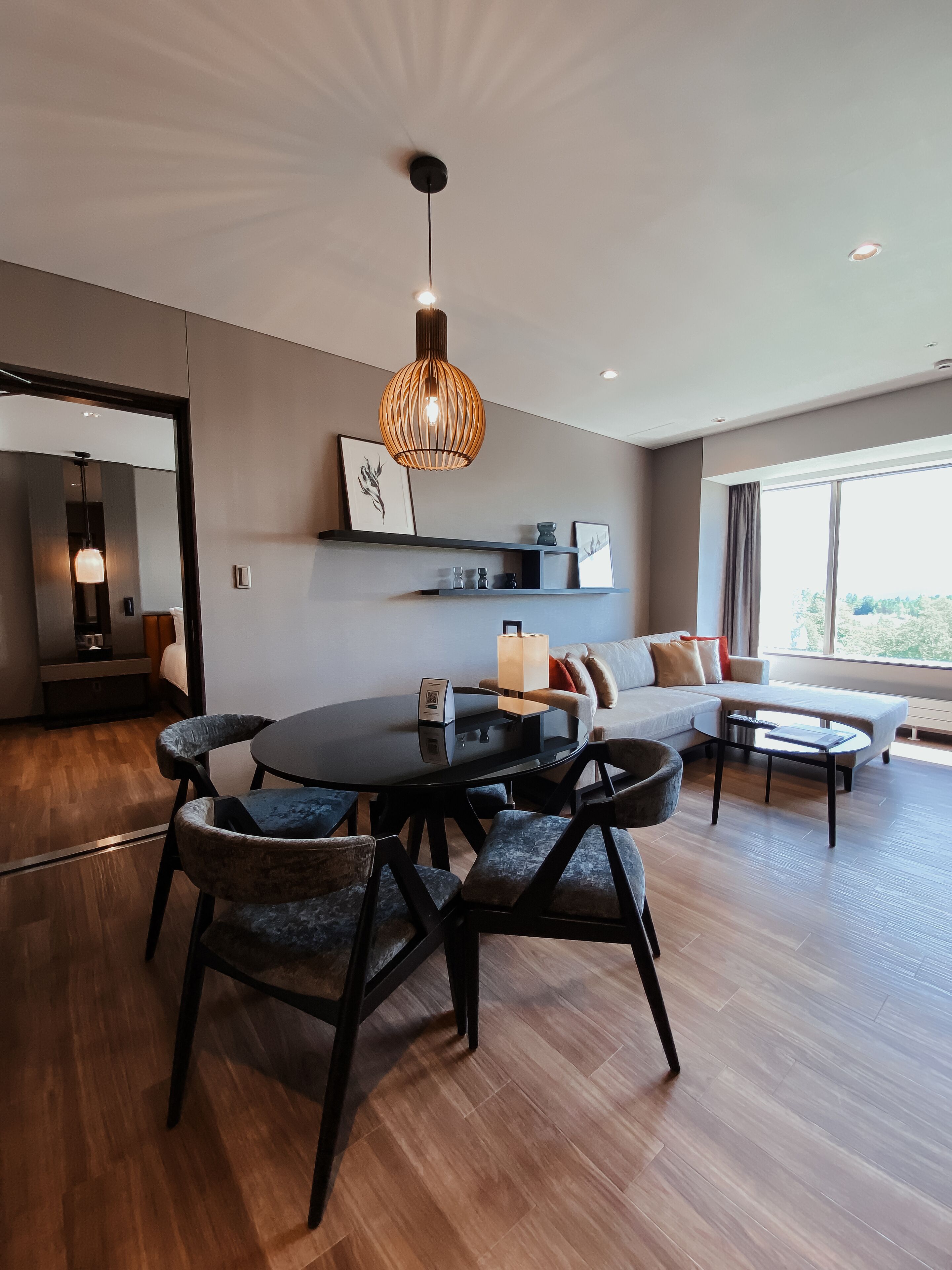 A modern living room with a round dining table, dark upholstered chairs, a sofa, and a large window offering a view.