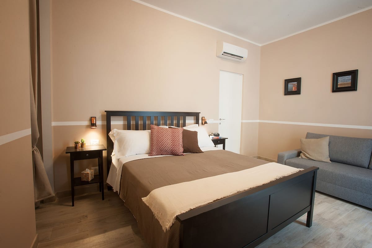 A bedroom with a bed, bedside tables, a nightstand, a sofa, and a window.