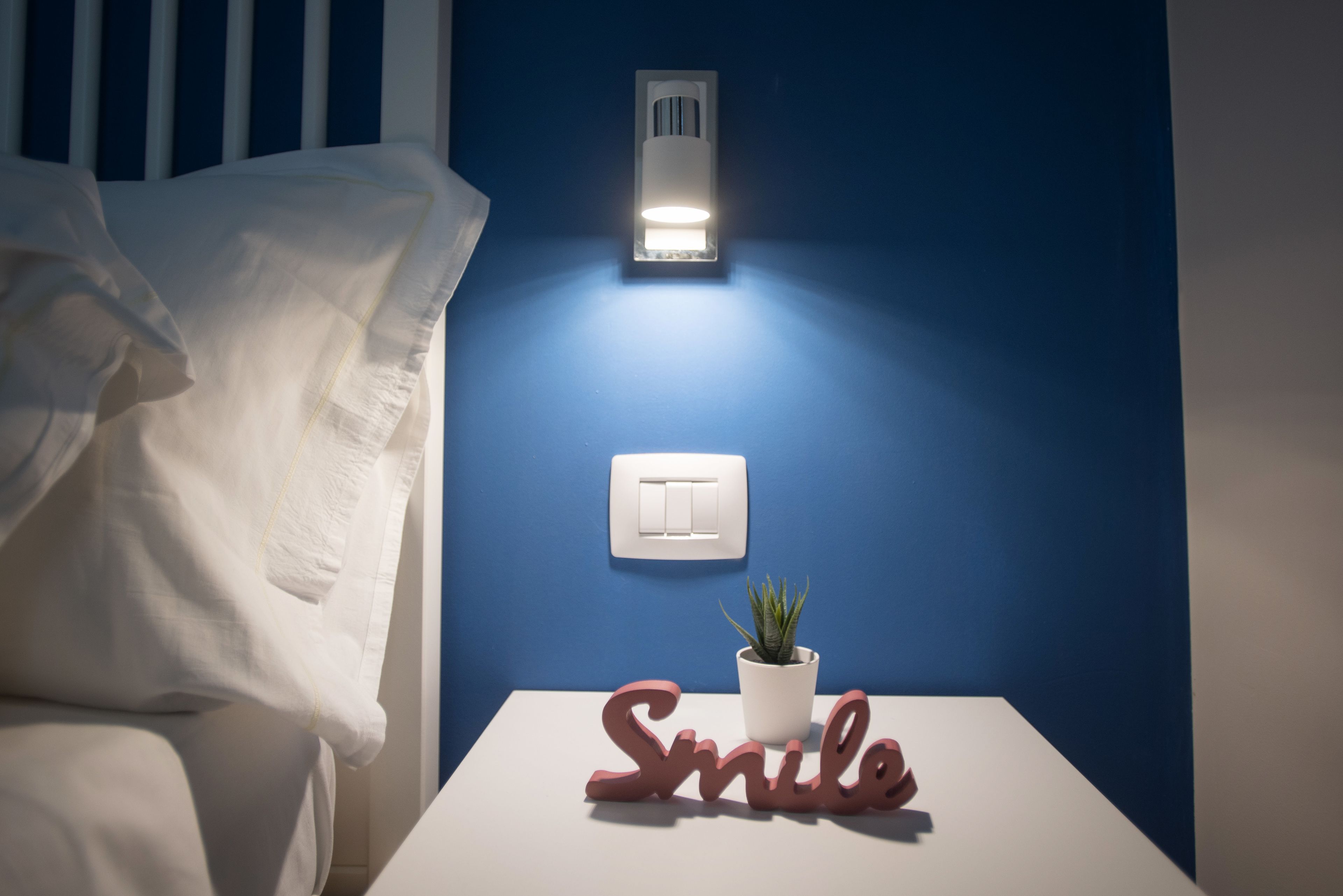 Ein Schlafzimmer mit einem Bett, einem Nachttisch mit Topfpflanze, einer Wandleuchte, einem Lichtschalter an der Wand und dem dekorativen Wort "Smile" auf dem Nachttisch.